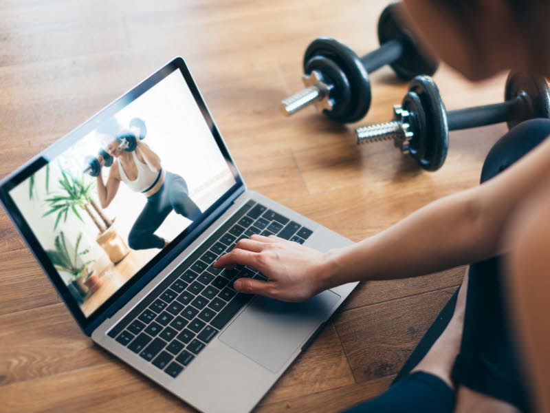 Illustrasjonsbilde av en PC som står på et gulv og en dame som trykker på tastaturet. Hun ser på en treningsvideo og det ligger håndvekter i bakgrunnen. Hun er klar for å trene på treningsrommet hun har laget seg hjemme.