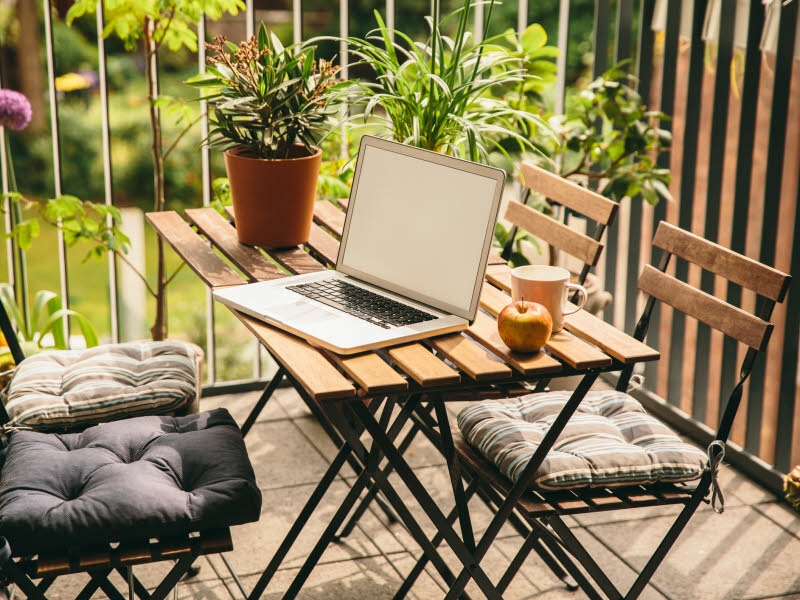Bildet viser en balkong med et lite brunt balkong bord og fire stoler med grå puter. Det står en laptop på bordet og viser hvordan man også kan bruke balkongen som hjemmekontor. Grønne planter vises i forgrunnen.