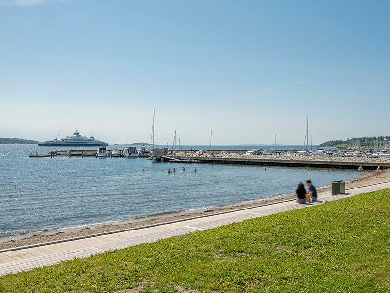 Bildet viser strandpromenaden i Moss. Grønt gress, sandstrand og blått hav som går i ett med den blå himmelen.