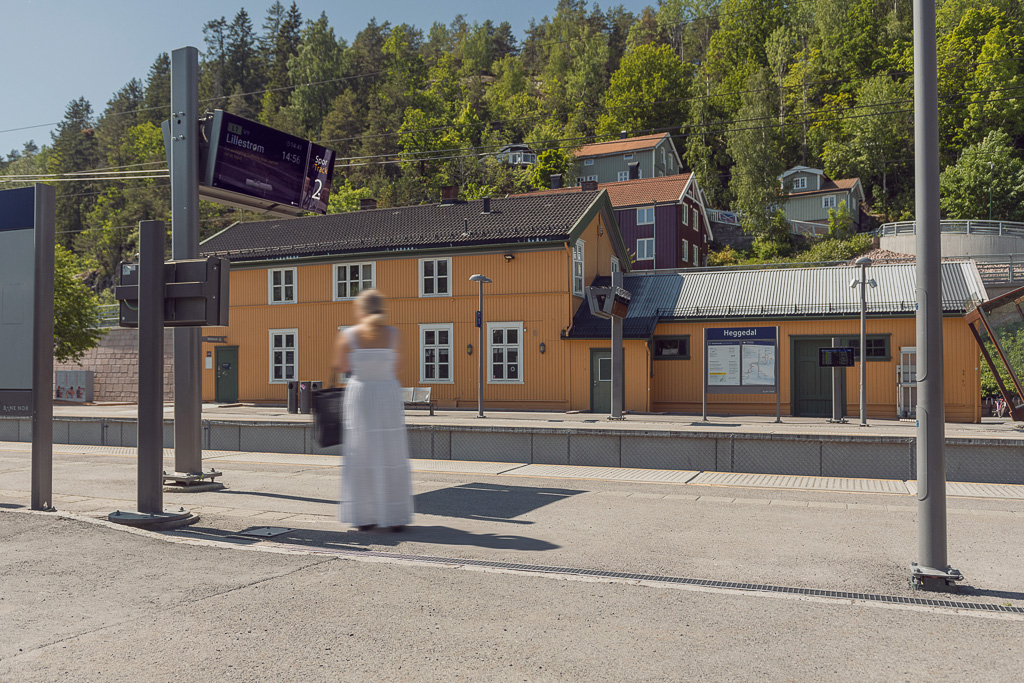Foto av togstasjonsområdet i Heggedal, som består av to perronger med et gult stasjonsbygg. En dame i hvit kjole står og venter på toget.