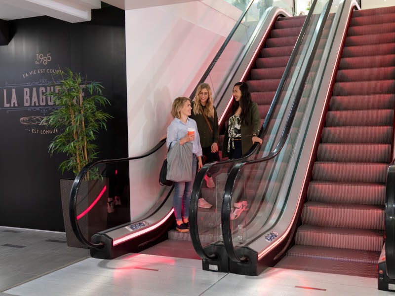 Bildet viser tre damer i en rulletrapp på et kjøpesenter. De er på shopping og har handleposer i hendene. 