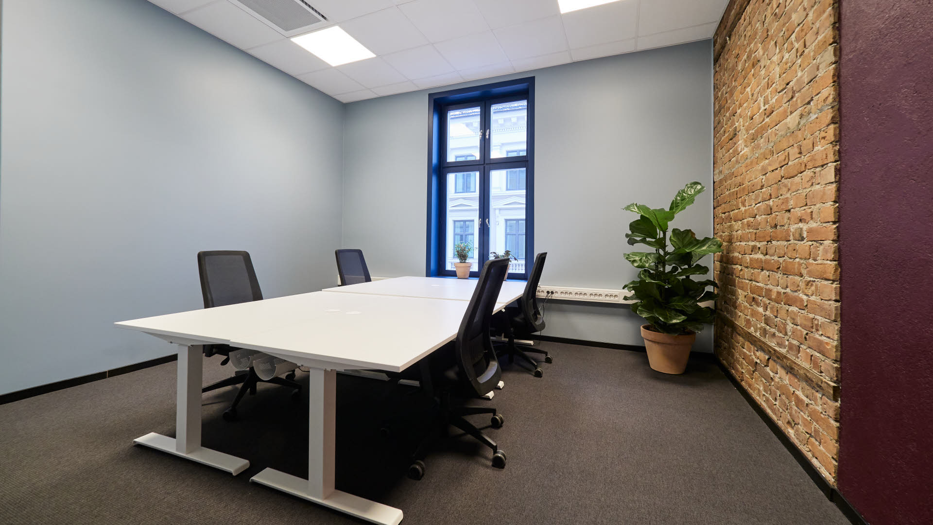 Bildet viser et kontorlokale i Thon Flex, Karl Johans gate 5. Den ene veggen har lys blå farge, ellers har veggene en dyp lilla farge med innslag av murstein. Hvite kontorpulter med svarte stoler. Et stort vindu gir mye lys i lokalet.