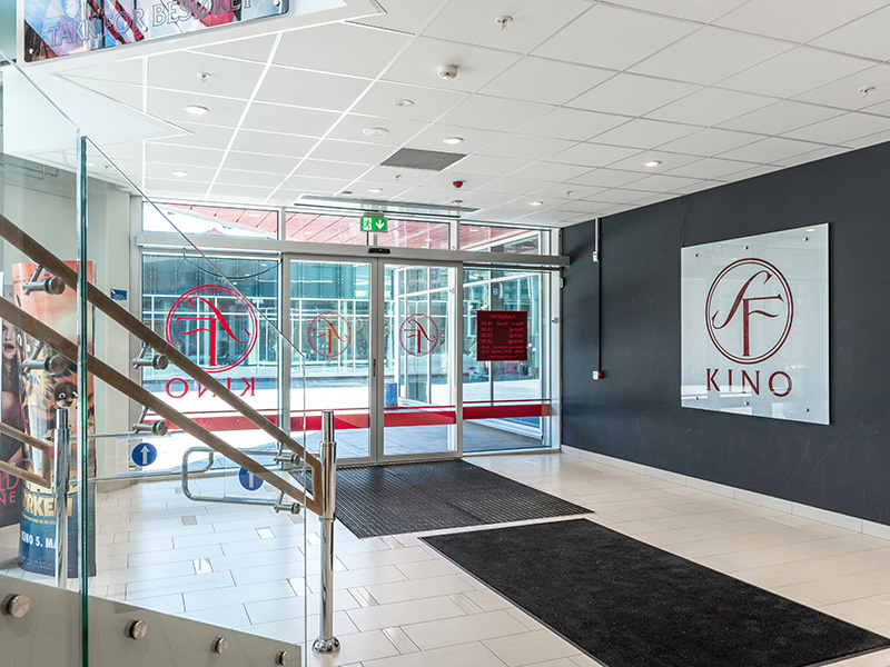 Bildet viser inngangen til kinoen på Sartor Storsenter. Svart teppe på gulvet, store glassdører og logoen til SF kino som bilde på en mørk grå vegg.